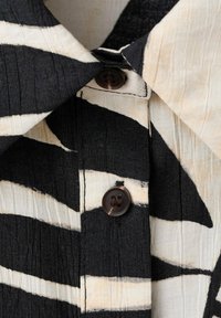 Gros plan d’un tissu rayé noir et beige avec deux boutons foncés et une couture visible au col d’un vêtement.