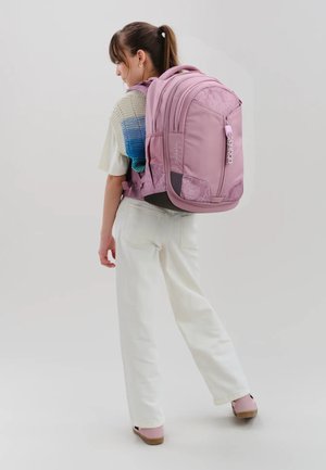 Young woman wearing a large pink backpack, white pants, and a multicolored knit top, standing sideways against a plain background.