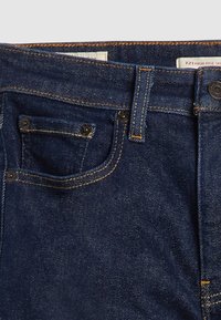 Jean en denim bleu foncé, taille haute, avec des coutures contrastantes, une petite poche avant et une fermeture à bouton. Texture lisse avec une coupe classique.