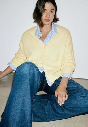 Femme aux cheveux courts et foncés assise par terre, portant un cardigan jaune sur une chemise bleue rayée et un jean large bleu sur fond blanc.