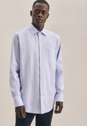 Jeune homme portant une chemise bleu clair à manches longues avec une poche poitrine, debout devant un fond clair uni.