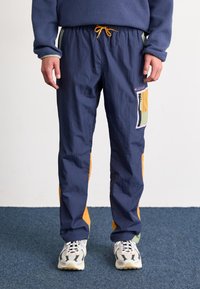 Pantalons légers bleus avec des accents oranges, featuring une poche latérale à motif, une taille élastique et une coupe décontractée. Tissu texturé.