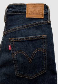 Σκούρο μπλε τζιν από denim με ταμπά δερμάτινο ταμπελάκι. Διαθέτει πορτοκαλί ραφές και κόκκινη ετικέτα στην πίσω τσέπη. Κλασικός σχεδιασμός με καμπυλωτές ραφές.