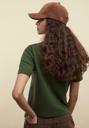 T-shirt vert à manches courtes avec un col rond, associé à une casquette en daim marron ; le dos du modèle est tourné, mettant en valeur des cheveux lâches et ondulés.