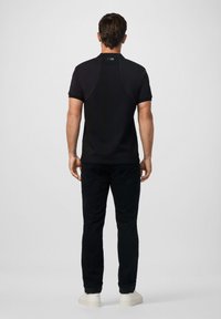 Muž v čiernom polo tričku a čiernych nohaviciach, stojaci chrbtom k jednému svetlému pozadiu.