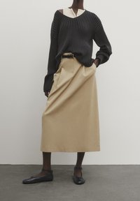 Personne portant un pull noir côtelé, une jupe midi beige avec ceinture et des ballerines noires à bride cheville, debout devant un fond clair uni.