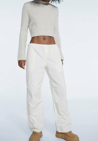 Hellbeige, gerippter, kurz geschnittener Pullover mit langen Ärmeln, kombiniert mit locker sitzenden, weißen Hosen, die einen elastischen Bund und eine Kordel haben.