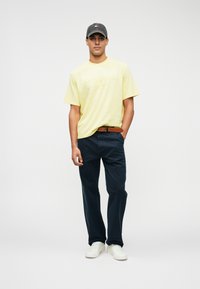 Gul bomull t-skjorte med preget tekst, marineblå bukser, brunt lærbelte og hvite joggesko; modell står foran en nøytral bakgrunn.