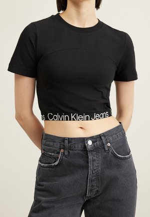 Frau trägt ein eng anliegendes schwarzes Crop-Top mit dem Calvin Klein Jeans Logo-Band, kombiniert mit hoch taillierten, ausgewaschenen schwarzen Jeans.