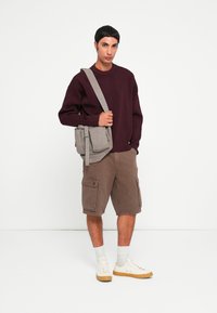 Sweatshirt tricoté bordeaux, short cargo beige, sac à bandoulière gris, baskets blanches avec semelles en caoutchouc naturel et chaussettes claires. Design simple et fonctionnel.