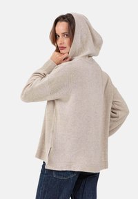 Beige gestrickte Kapuzenpullover mit weicher Textur, die eine lockere Passform und gerippte Bündchen aufweist. Seitliche Schlitze verleihen dem Design zusätzliche Details.