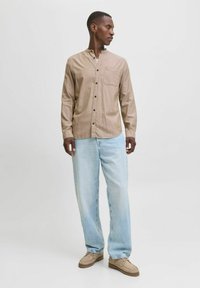 Light brown button-up shirt with a pocket, paired with light blue jeans and beige shoes. Textured fabric with a relaxed fit.