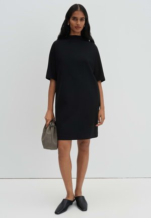 Vrouw staat in een zwarte jurk met korte mouwen, houdt een grijze handtas vast en draagt zwarte instapschoenen tegen een eenvoudige lichte achtergrond.