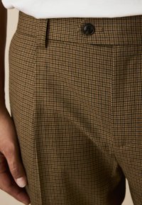 Brown plaid trousers with a textured fabric, featuring a black button and a tailored waistband. Side pocket partially visible.