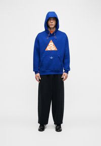 Άνδρας με μπλε φούτερ με κουκούλα και πορτοκαλί λογότυπο ACG, μαύρο φαρδύ παντελόνι και μαύρα λαμπερά παπούτσια, στέκεται μπροστά σε λευκό φόντο.