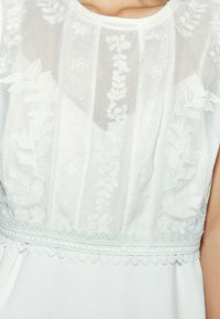 Robe blanche avec un corsage transparent brodé, motifs floraux et dentelle le long du décolleté et de l'ourlet. Texture de tissu doux et léger.