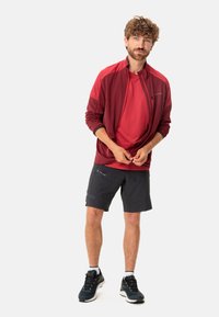 Giacca leggera color marsala sopra una maglietta rossa, abbinata a pantaloni corti neri e scarpe da trekking blu. Presenta una chiusura con zip anteriore e polsini elasticizzati.