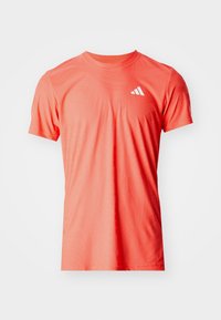 Koszulka sportowa z krótkim rękawem w jasnym koralowym kolorze, z teksturowanym wzorem i białym logo na górnej lewej stronie. Wykonana z oddychającego materiału.
