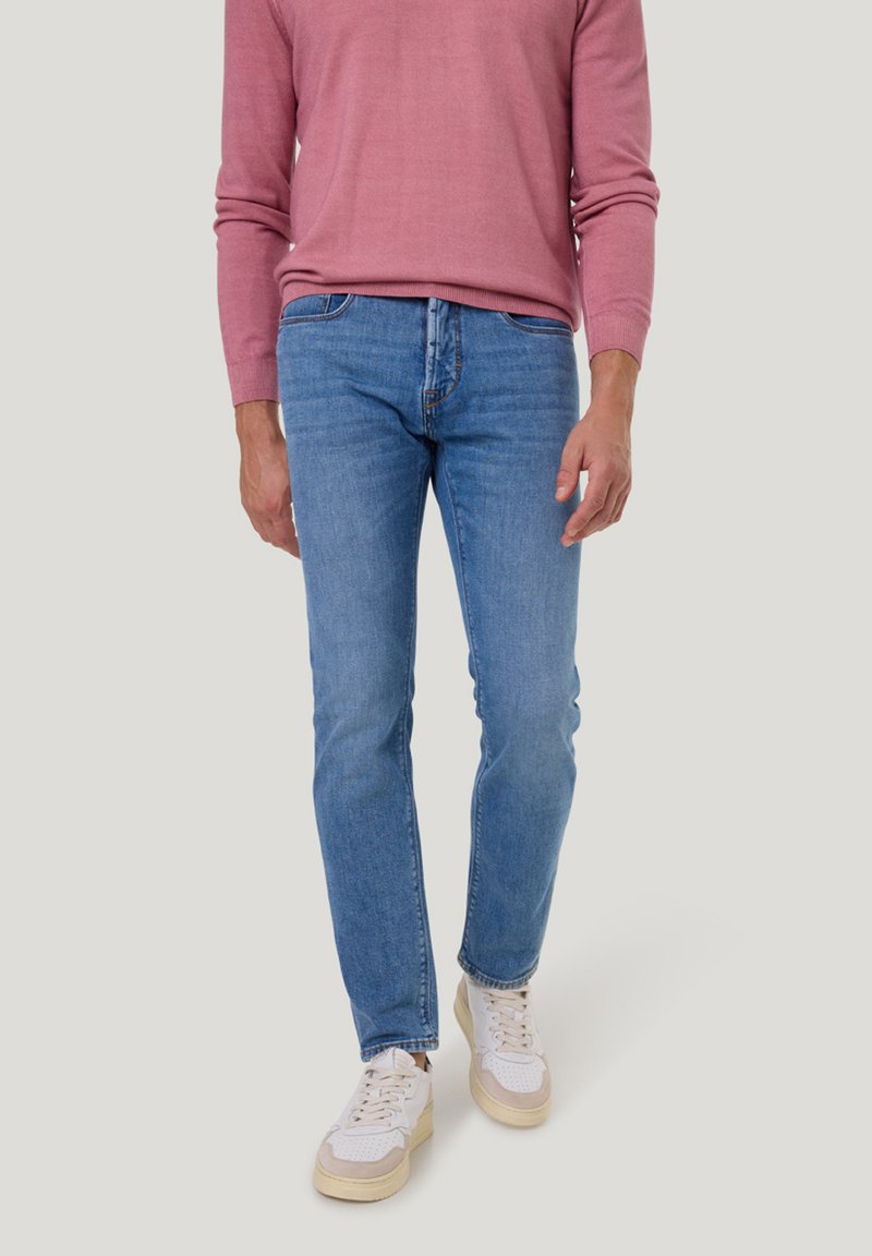Helle blaue Herrenjeans in Slim-Fit, aus Denim gefertigt. Kombiniert mit einem rosa Langarmhemd und weißen Sneakern mit beigen Akzenten.
