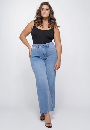 Vrouw met golvend haar, gekleed in een zwart mouwloos topje, lichtblauwe jeans met hoge taille en zwarte sandalen met bandjes, poserend tegen een effen achtergrond.