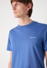 Blue cotton t-shirt features a round neck and short sleeves, with a white "Calvin Klein" logo on the left chest. Smooth texture.