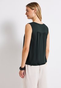 Tmavě zelený bezrukávový top se skládáním na zadní straně, hladkým texture a elastickým lemem. Slučuje se s světlými, volně střiženými kalhotami.