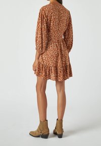 Donna che indossa un abito floreale marrone con lunghe maniche a sbuffo e orlo arricciato, abbinato a stivali alla caviglia in suede beige, in piedi rivolta di spalle.