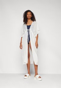 Kimono trasparente bianco con motivo a spina di pesce, caratterizzato da maniche ampie e una chiusura frontale, indossato sopra un costume da bagno blu navy e sandali con piattaforma.