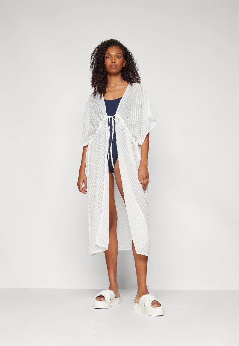Kimono trasparente bianco con motivo a spina di pesce, caratterizzato da maniche ampie e una chiusura frontale, indossato sopra un costume da bagno blu navy e sandali con piattaforma.