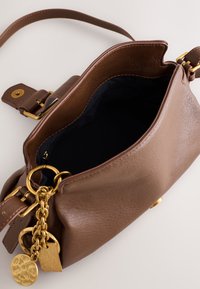 Bruine leren handtas met gouden ketting en bedels, open bovenkant die de zwarte binnenvoering en een ritsvak laat zien, op een witte achtergrond.