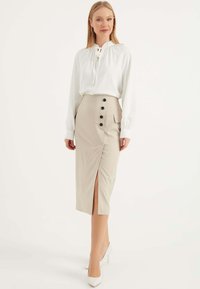 Weiße Bluse mit gerafftem Ausschnitt, kombiniert mit einem beigen Bleistiftrock mit Schlitz und schwarzen Knopf-Akzenten entlang der Taille. Weiße High Heels.