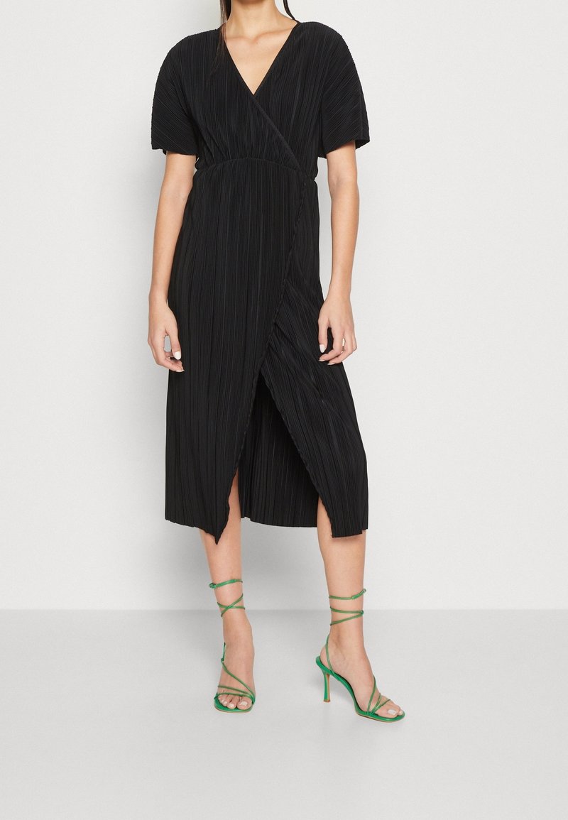 Femme portant une robe midi plissée noire à manches courtes et des sandales vertes à talons avec brides, debout devant un fond blanc uni.
