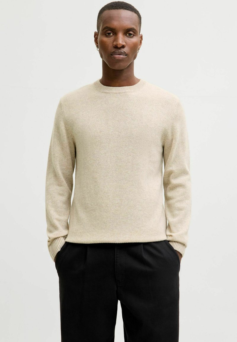 Beige kasjmier trui met een ronde halslijn, lange mouwen en geribbelde manchetten, gecombineerd met zwarte broeken. Gladde textuur, minimalistisch ontwerp.