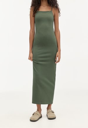 Femme portant une robe ajustée sans manches, longue jusqu’aux chevilles, de couleur vert olive, avec des chaussures beige à enfiler avec des boucles, debout devant un fond uni.