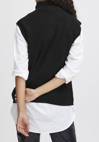 Černý žebrovaný bezrukávový svetr s vysokým límcem, nošený přes bílou košili s dlouhým rukávem, který ukazuje vrstvený vzhled a jemnou strukturu.