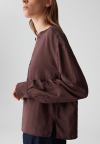 Woman wearing a loose, long-sleeved, dark brown shirt with cuff buttons and side slits, shown from the side against a plain background.