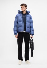 Giacca imbottita blu con colletto alto, giacca nera con zip sotto, pantaloni neri e sneaker color crema. Tiene una borsa nera con tracolla.