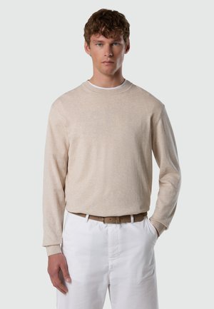 Junger Mann trägt einen beigen langärmeligen Pullover, der in weiße Hosen mit einem braunen Gürtel gesteckt ist, steht mit einer Hand in der Tasche vor einem einfarbigen Hintergrund.