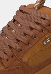 Gros plan sur une basket en daim marron avec des lacets épais, des détails de couture et un petit patch logo rectangulaire sur le côté.