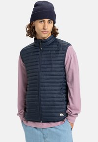 Marineblaue Steppweste mit hohem Kragen und horizontalem Steppmuster, kombiniert mit einem hellrosa Langarmshirt und blauen Hosen.