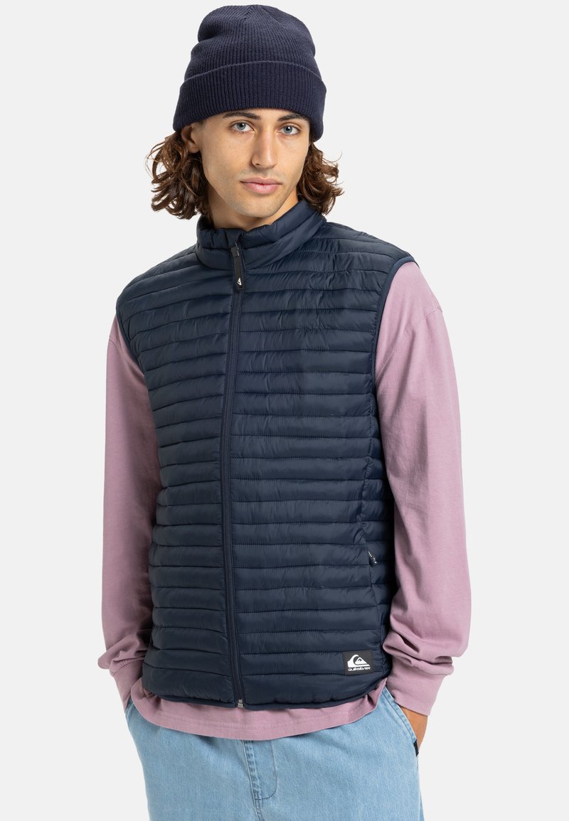 Marineblaue Steppweste mit hohem Kragen und horizontalem Steppmuster, kombiniert mit einem hellrosa Langarmshirt und blauen Hosen.