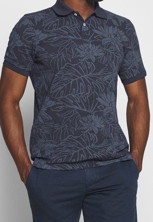 Homme portant un polo bleu marine avec des motifs floraux et de feuilles bleu clair, associé à un pantalon bleu foncé, les mains dans les poches.