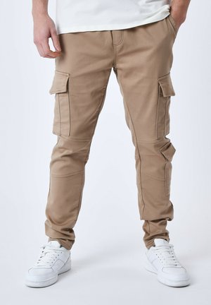 Pantalones cargo beige con múltiples bolsillos laterales, piernas ajustadas y costuras reforzadas. Llevados con zapatillas blancas.