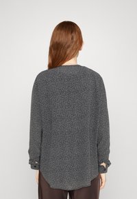 Blusa nera a maniche lunghe con piccole macchie bianche, caratterizzata da un orlo ondulato e polsini con bottoni. Realizzata in un tessuto leggero e testurizzato.