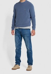 Pull bleu avec poignets et ourlet côtelés, porté sur une chemise grise. Assorti à un jean bleu et des baskets en daim beige. Tenue simple et décontractée.