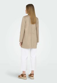 Donna con capelli lisci castano chiaro, che indossa un cappotto beige, pantaloni bianchi a gamba larga e sandali con zeppa marroni, in piedi di spalle su uno sfondo semplice.