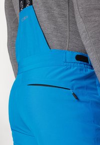 Salopette bleue imperméable avec bretelles noires ajustables, poche zippée à l'avant et texture lisse, conçue pour les activités en plein air.