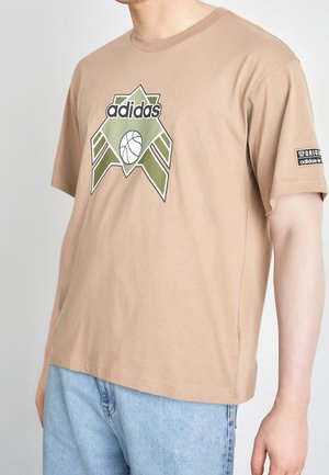 Beiges Adidas T-Shirt mit grünem geometrischem Logo und Basketballgrafik vorne, getragen von einer Person in hellblauen Jeans.