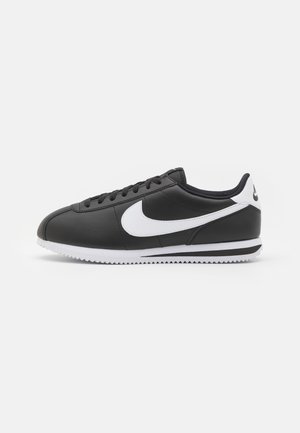 Zapatilla de cuero negro con un swoosh y acentos en blanco. Presenta una punta redondeada, cierre con cordones y una suela de goma blanca texturizada.
