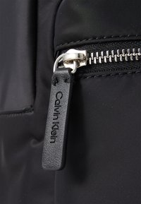 Mochila negra de nailon con cremallera plateada y etiqueta de cuero. La etiqueta tiene "Calvin Klein" grabado. Textura suave, diseño minimalista, hardware duradero.